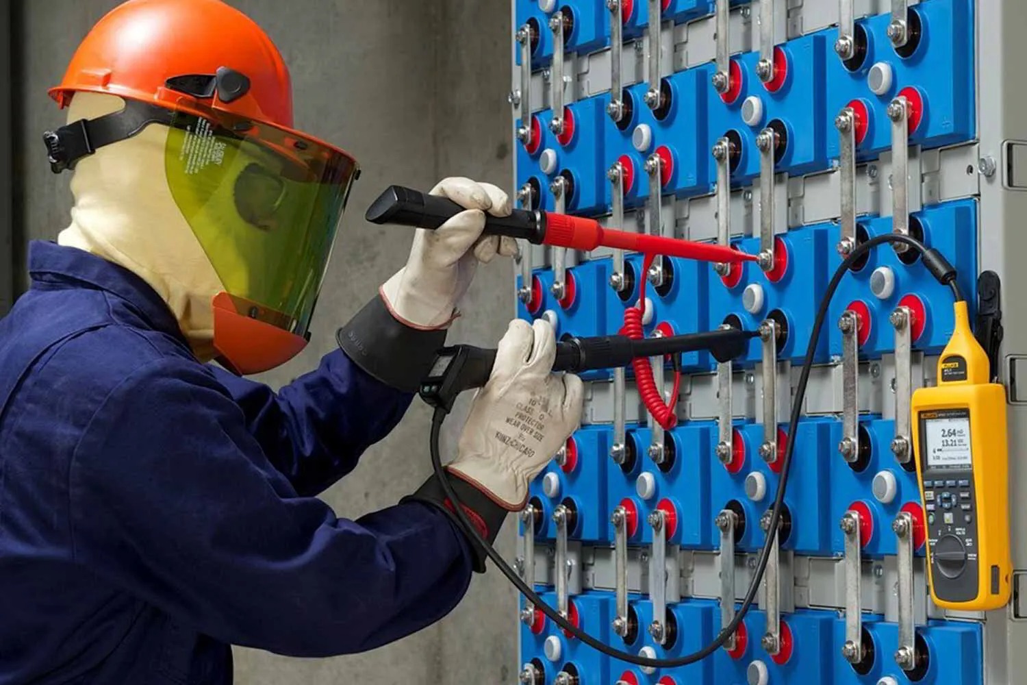 Worker in protective gear tests a battery bank using probes and a handheld meter.