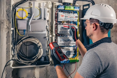Electrician wearing safety gear uses a multimeter on an open electrical panel.